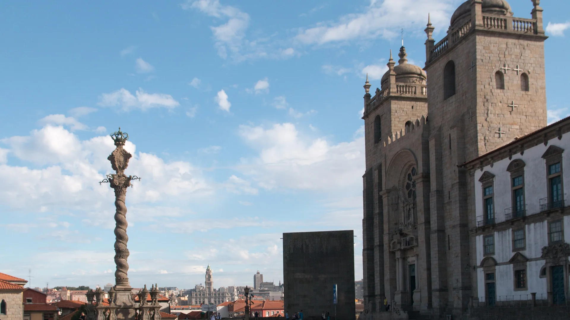 Porto Cathedral and its square under a blue sky, with the stone pillory column on the left and the cathedral's twin towers on the right.