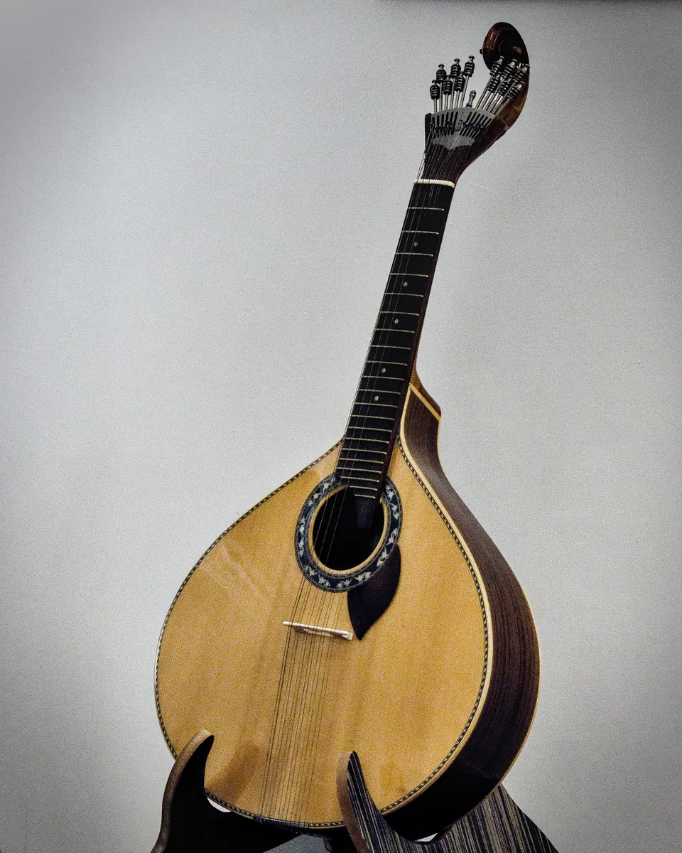 A single Portuguese guitar with a pear-shaped golden body and a scroll at the head, on a stand against a plain light wall.