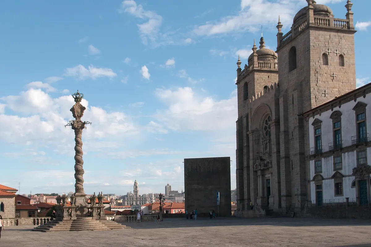 Porto Cathedral and its square under a blue sky, with a stone pillory column and the cathedral's twin towers.