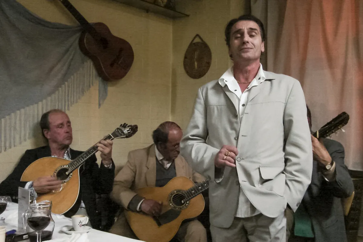 A fado singer in a light suit standing beside two seated guitar players in a small room in Porto.